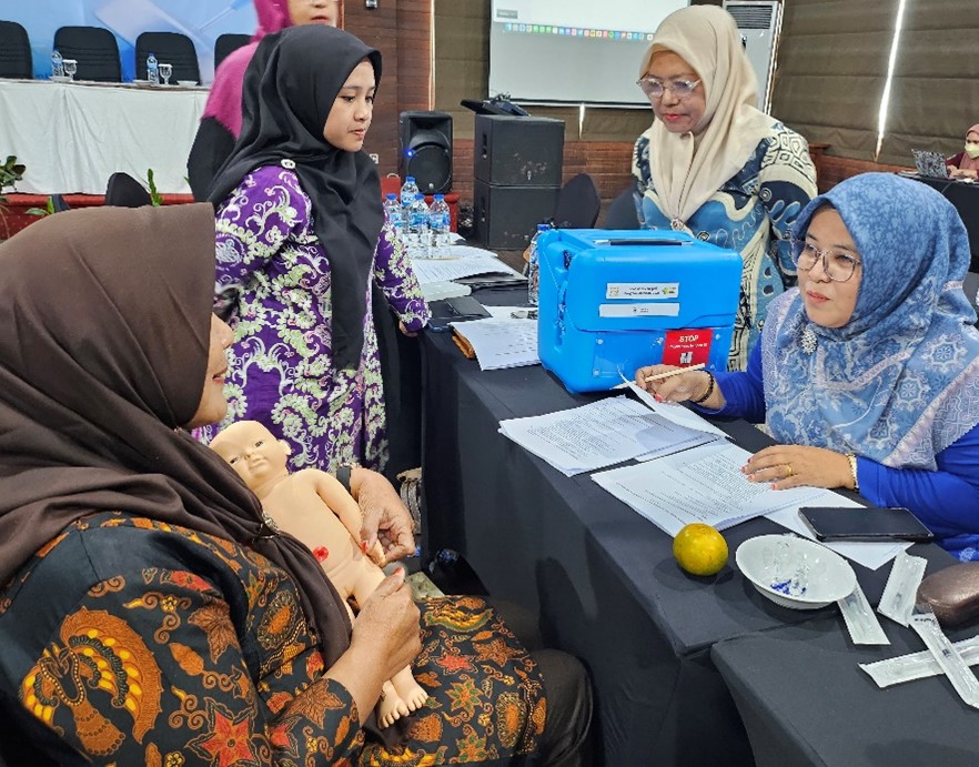 A woman hold a baby doll and two others act as health care workers in Posyandu. A training instructor oversees the process.